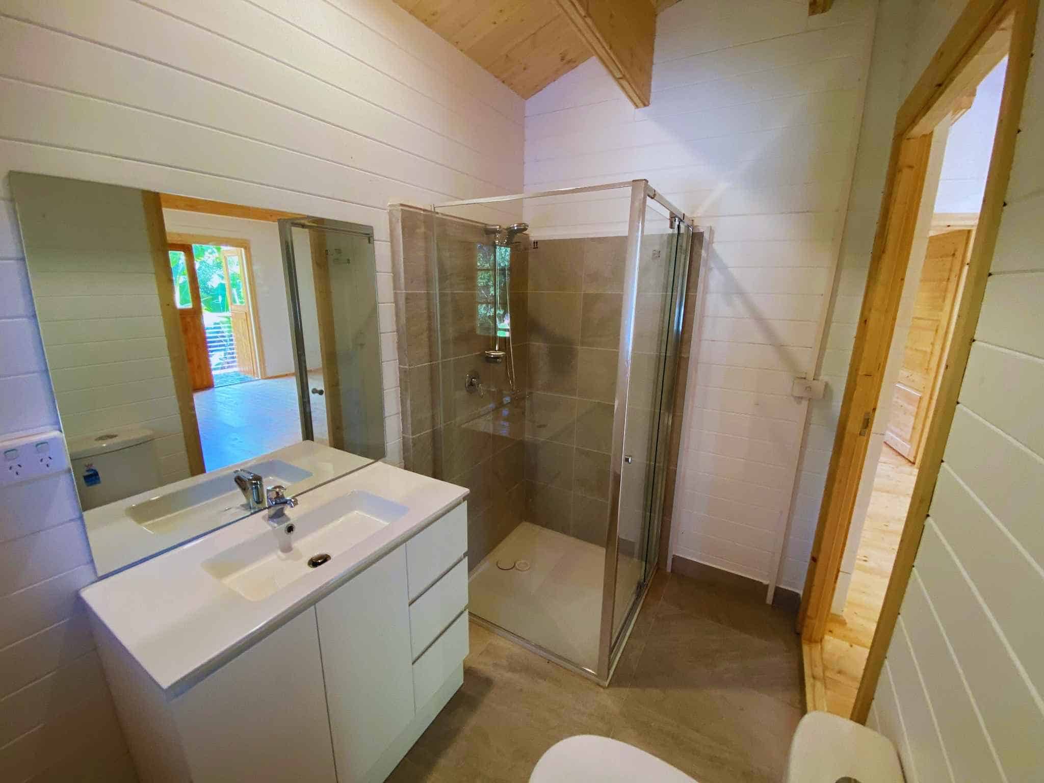 Modern bathroom featuring a white vanity with a sink, a large mirror, and a glass shower with beige tiles, set against a light wood interior.