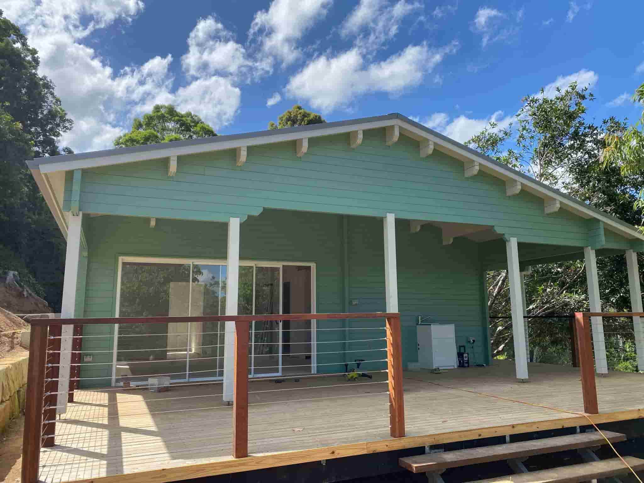 Granny flat with a light green exterior, wooden deck with modern wire railing, and large glass doors, surrounded by lush greenery and blue skies.