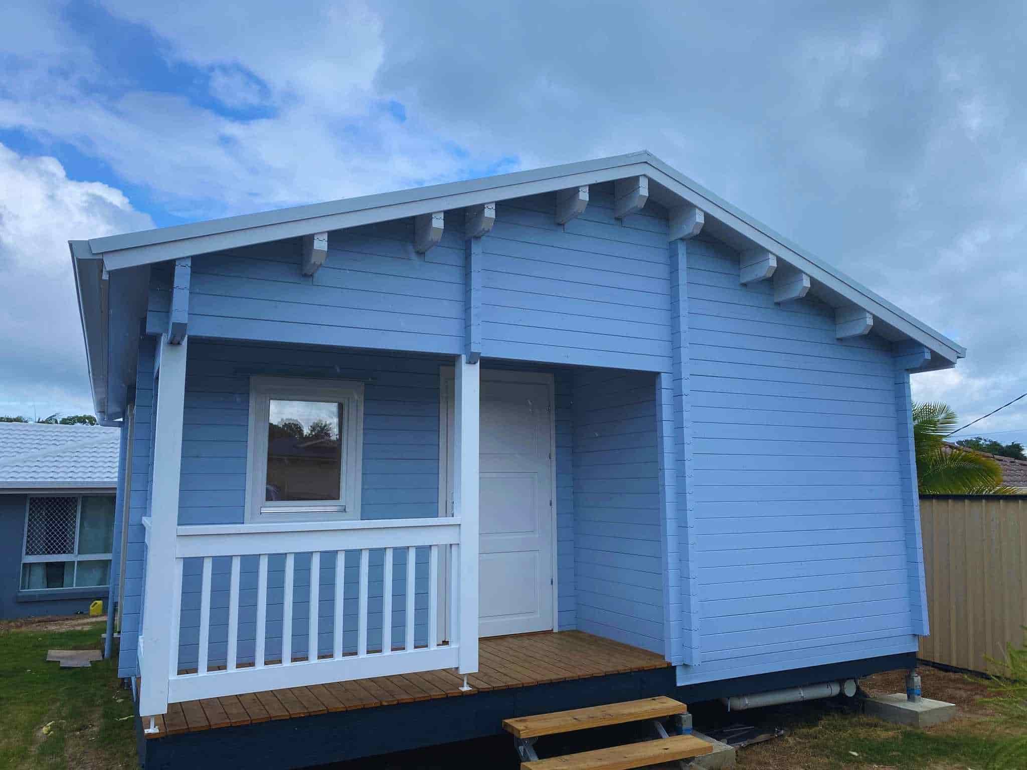 Granny flat with a soft blue exterior, white railing on the front porch, and wooden steps, set against a partly cloudy sky.