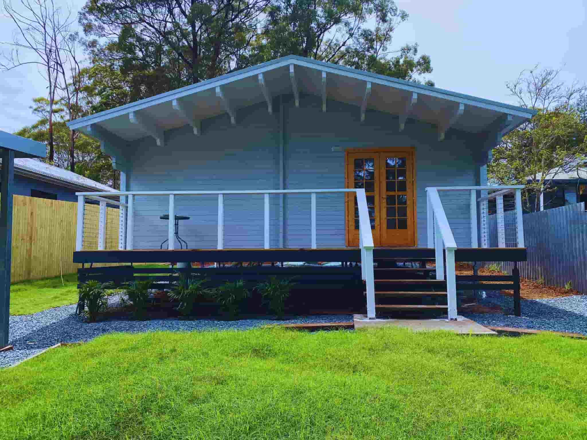 Granny flat with a light blue exterior, wooden deck, white railing, and a set of double French doors, surrounded by gravel pathway and green lawn.