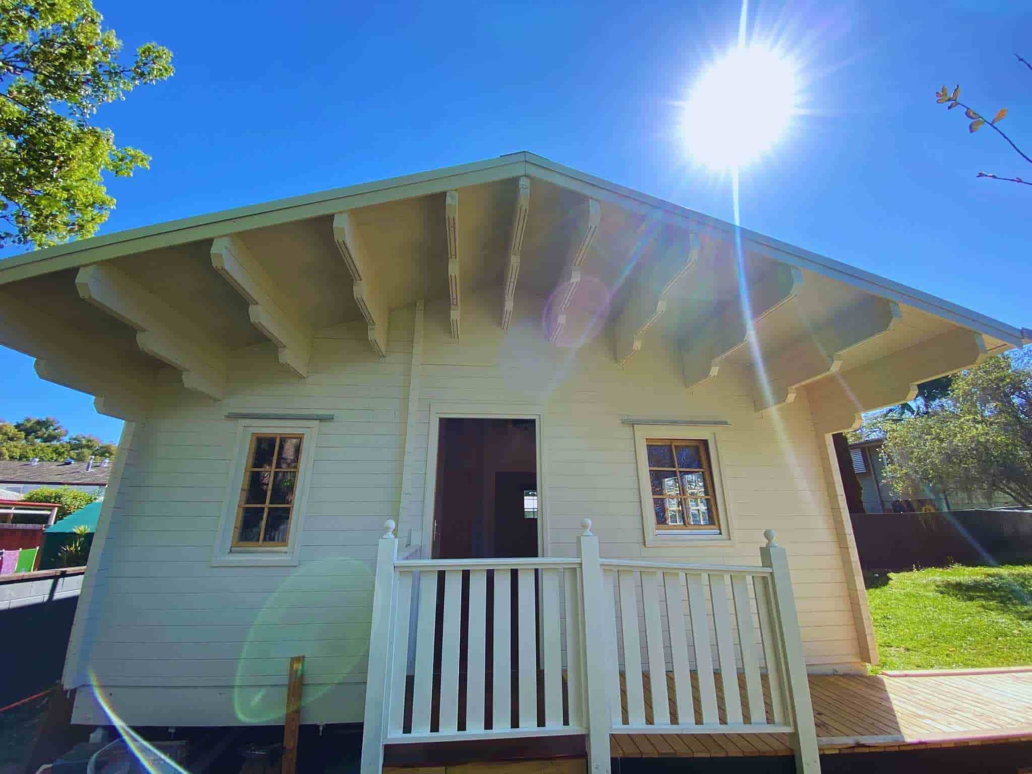 Granny flat with a light beige exterior, front porch with white railing, and large sunlit windows, set against a clear blue sky.