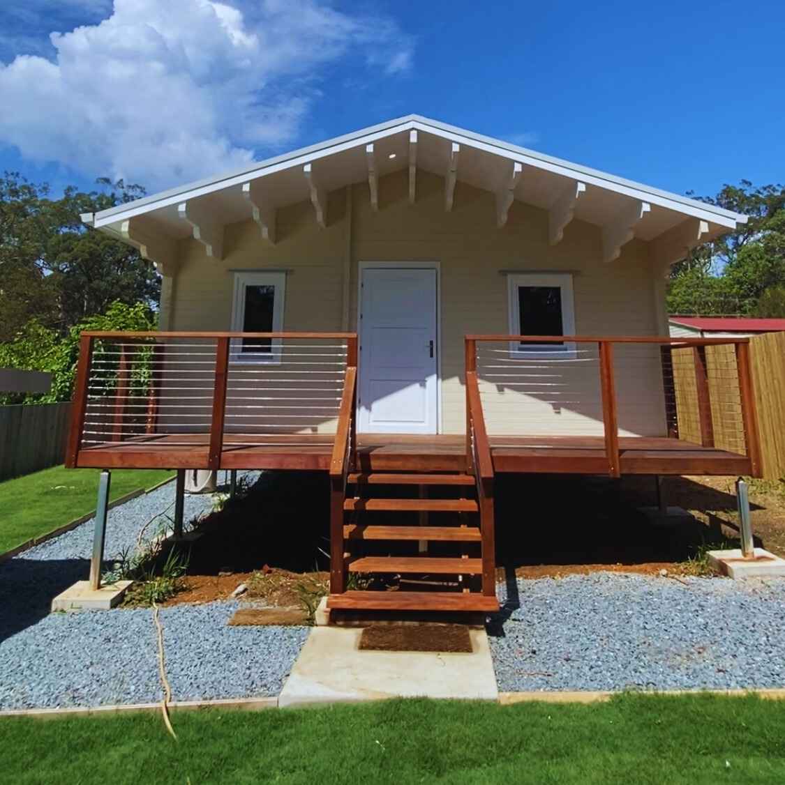 Granny flat with a light beige exterior, wooden deck with modern wire railing, front staircase, and landscaped yard with gravel and green grass.