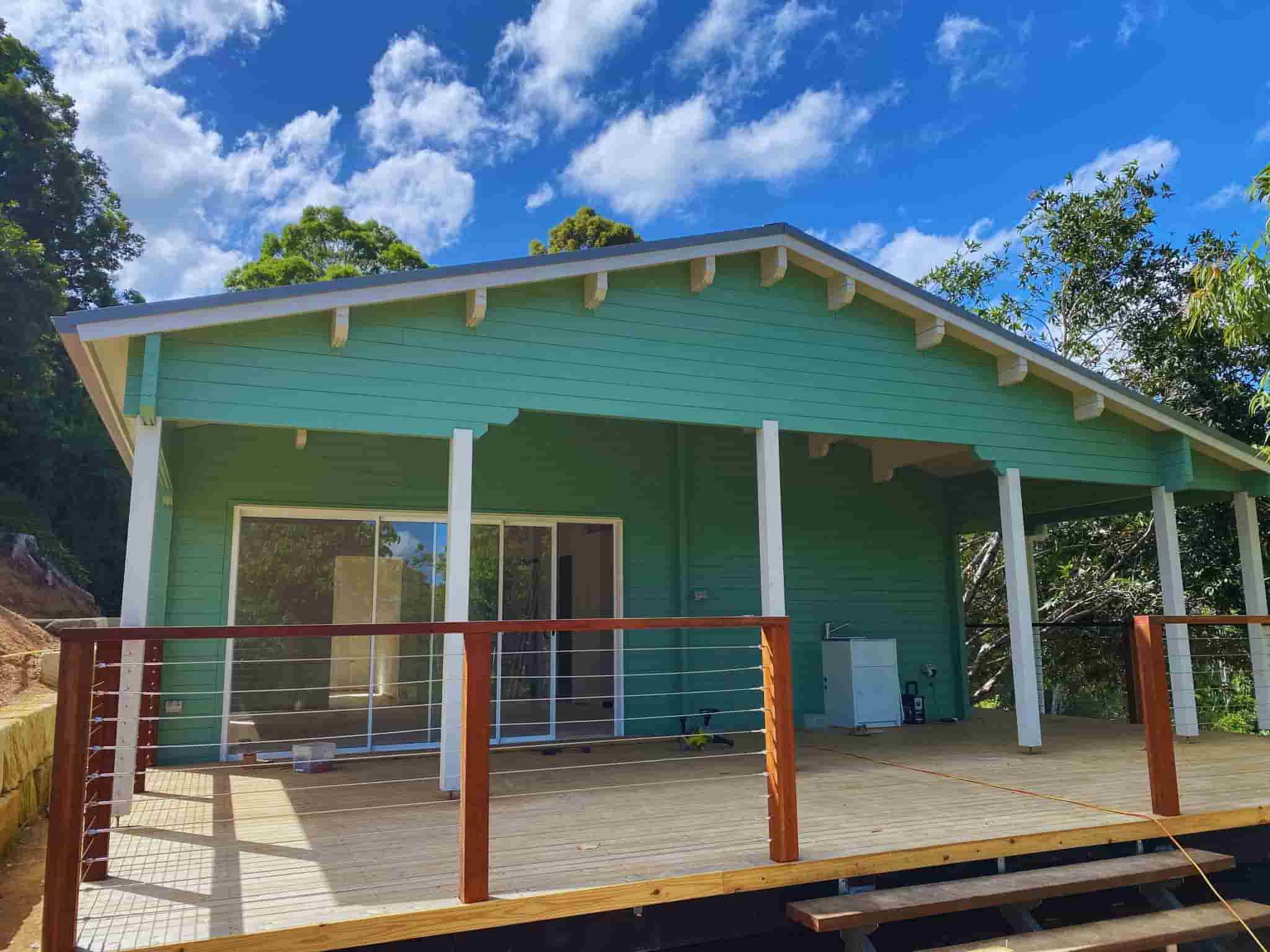 Modern granny flat with a soft green exterior, spacious wooden deck, and sleek railing, surrounded by trees and clear skies.