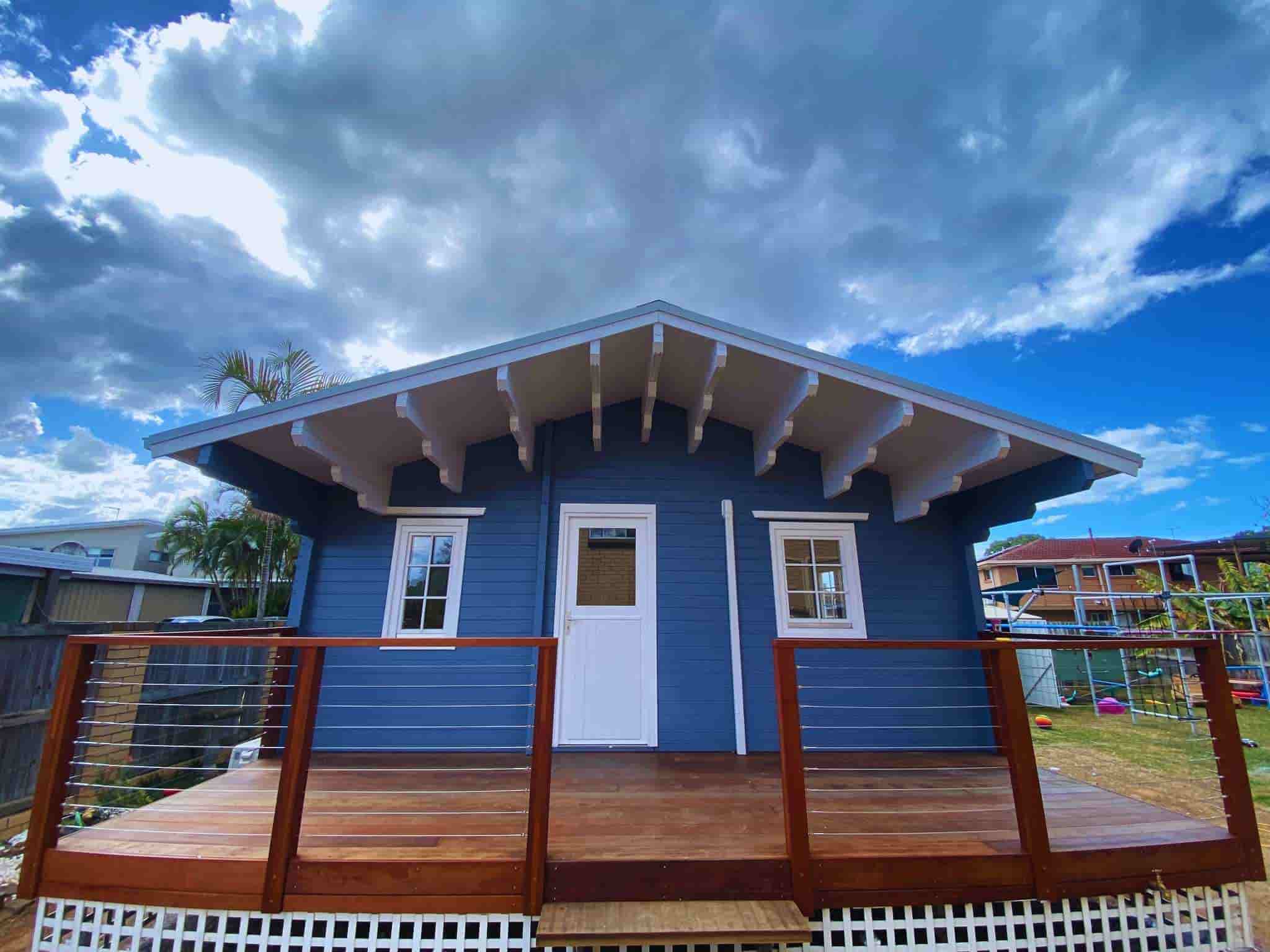 Granny flat with a deep blue exterior, wooden deck with modern wire railing, and large windows, set against a dramatic sky with clouds.