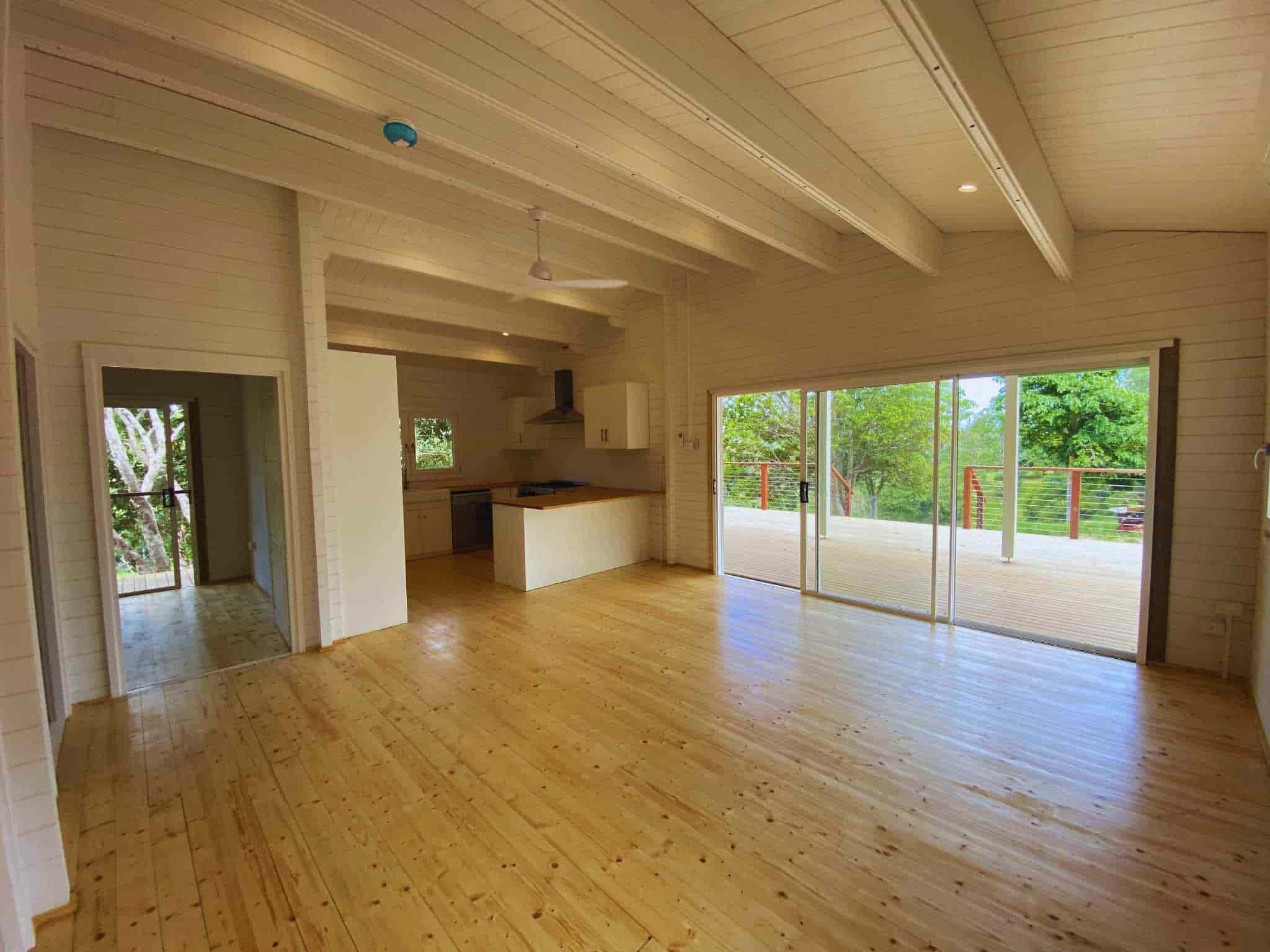 Open-plan living area of a granny flat with high ceilings, wooden floors, a kitchen with white cabinets, and large sliding doors leading to a spacious deck and natural surroundings.