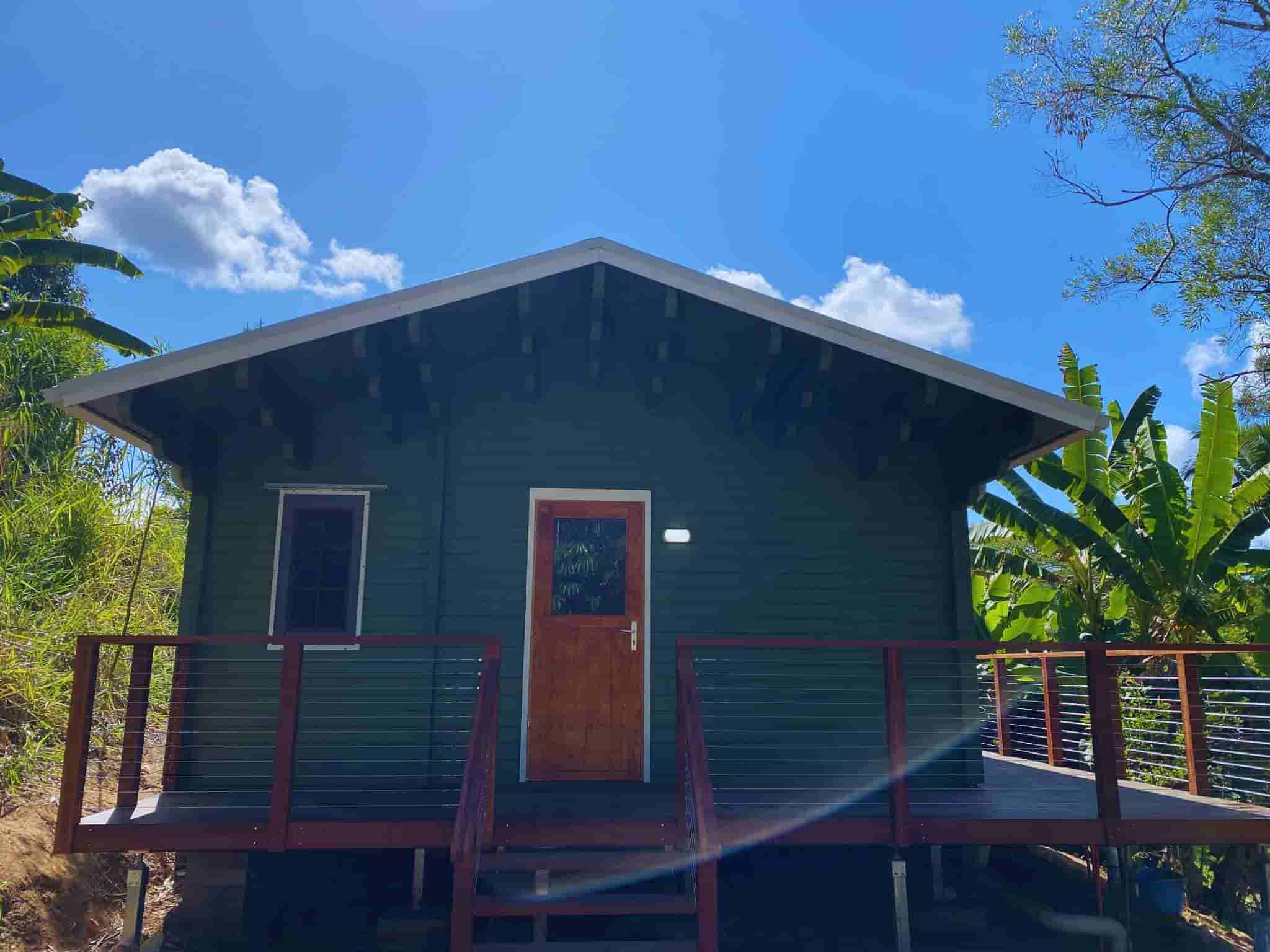 Granny flat with a dark green exterior, wooden deck with modern wire railing, and a wooden front door, surrounded by lush greenery and bright blue skies.