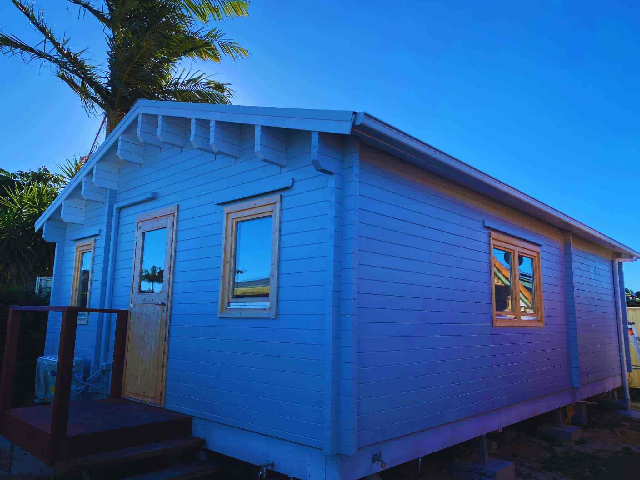 Side view of a light blue granny flat with wooden door and windows, small front step, and surrounded by bright blue skies and greenery.