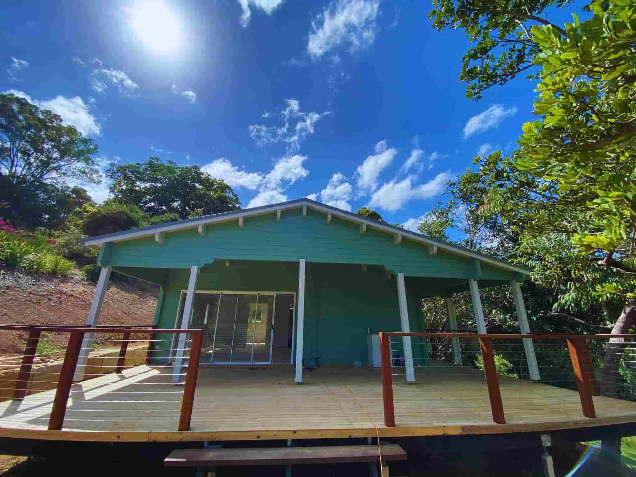 Granny flat with a light green exterior, large wooden deck with modern wire railing, and floor-to-ceiling glass doors, surrounded by greenery and bright blue skies.
