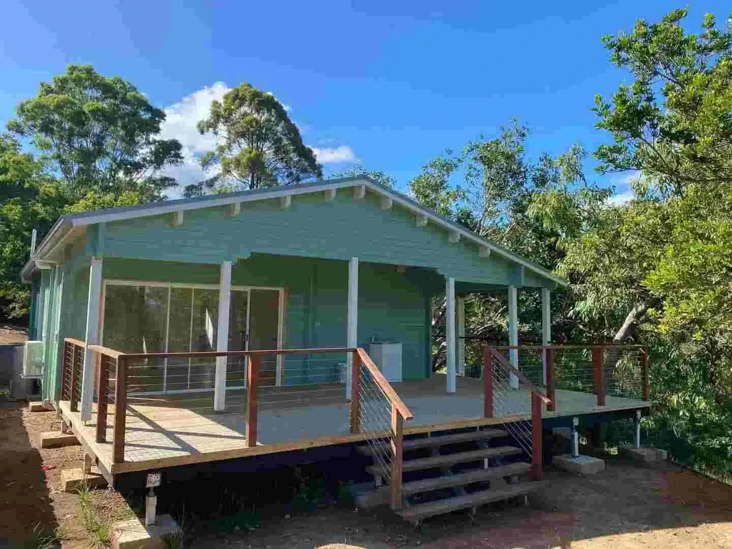 Charming granny flat with a soft green facade, spacious wooden deck, large glass doors, and a scenic backdrop of trees.